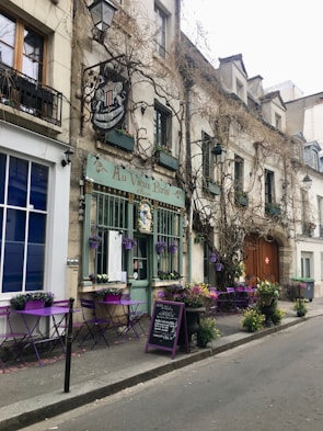 A charming Parisian street café with vintage appeal features purple tables and chairs aligned outside. Climbing vines decorate the building's exterior, and colorful flowers add a welcoming touch.