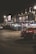 A dimly lit street scene at night featuring storefronts with signage including 'Smoke Alley' which advertises products such as fine glass, vaporizers, and e-cigarettes. A red car is parked in front of these shops, and the area appears quiet and empty.