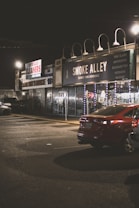 A dimly lit street scene at night featuring storefronts with signage including 'Smoke Alley' which advertises products such as fine glass, vaporizers, and e-cigarettes. A red car is parked in front of these shops, and the area appears quiet and empty.