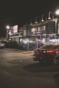 A dimly lit street scene at night featuring storefronts with signage including 'Smoke Alley' which advertises products such as fine glass, vaporizers, and e-cigarettes. A red car is parked in front of these shops, and the area appears quiet and empty.