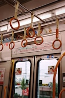 Inside a public transportation vehicle featuring orange hanging straps attached to a metallic rail. The interior includes informational posters and advertisements above the doors, with images likely showcasing products or services. Exterior elements such as trees and buildings are visible through the windows, suggesting motion through an urban area.