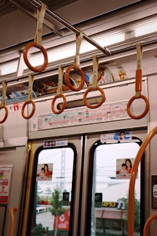 Inside a public transportation vehicle featuring orange hanging straps attached to a metallic rail. The interior includes informational posters and advertisements above the doors, with images likely showcasing products or services. Exterior elements such as trees and buildings are visible through the windows, suggesting motion through an urban area.