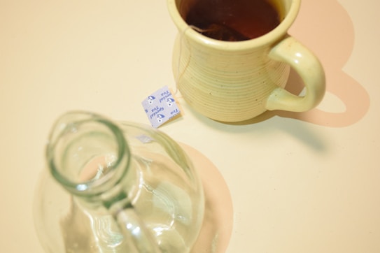 A cream-colored ceramic mug partially filled with tea, accompanied by a transparent glass bottle, is set on a light-colored surface. A small packet of sweetener rests beside the mug.