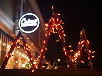 A row of red-orange Christmas lights is wrapped around a triangular frame, illuminating the dark night. In the background, there is a building with a neon circular sign that appears to be for a cafe or restaurant. Other lights from the building add a warm and inviting glow to the scene.