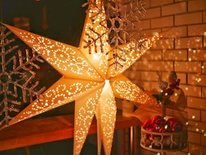 A large illuminated paper star lantern with intricate cut-out patterns emits a warm glow in a cozy setting. Decorative snowflakes hang nearby, adding a festive touch, while a small arrangement of red ornaments and a golden bow sits on a shelf below, enhancing the holiday ambiance.
