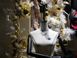 A mannequin is adorned with jewelry, including a necklace and brooch, set against a festive holiday backdrop. The scene is decorated with golden baubles, white garlands, and wrapped gifts with white bows, creating a luxurious and celebratory atmosphere.