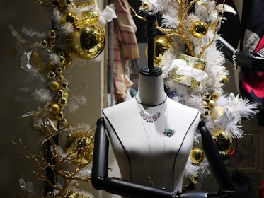 A mannequin is adorned with jewelry, including a necklace and brooch, set against a festive holiday backdrop. The scene is decorated with golden baubles, white garlands, and wrapped gifts with white bows, creating a luxurious and celebratory atmosphere.