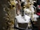 A mannequin is adorned with jewelry, including a necklace and brooch, set against a festive holiday backdrop. The scene is decorated with golden baubles, white garlands, and wrapped gifts with white bows, creating a luxurious and celebratory atmosphere.
