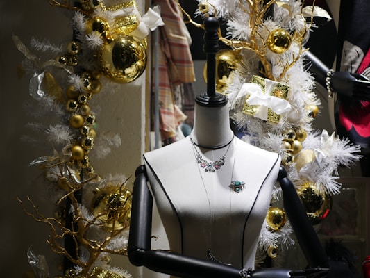 A mannequin is adorned with jewelry, including a necklace and brooch, set against a festive holiday backdrop. The scene is decorated with golden baubles, white garlands, and wrapped gifts with white bows, creating a luxurious and celebratory atmosphere.