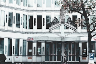 A modern, multi-story building with numerous windows, featuring three statues above the entrance. The entrance has glass doors and a sign with red letters above. A nearby tree with autumn leaves adds a touch of nature.
