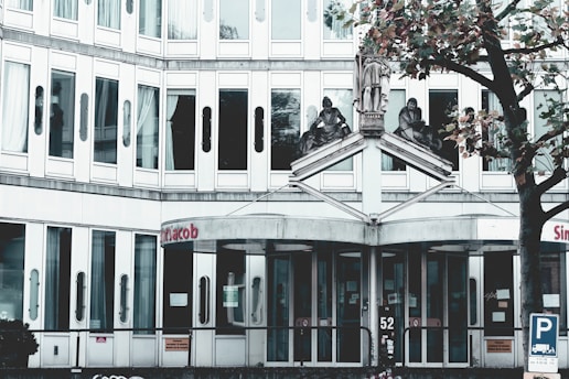 A modern, multi-story building with numerous windows, featuring three statues above the entrance. The entrance has glass doors and a sign with red letters above. A nearby tree with autumn leaves adds a touch of nature.