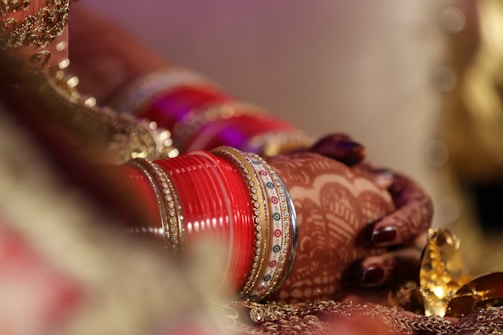 A close-up of a person's hands adorned with intricate henna designs and multiple red bangles. The scene is richly decorated with golden details, giving a sense of elegance and tradition.