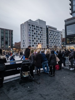 A bustling urban scene featuring an outdoor ice skating rink surrounded by modern buildings. People are ice skating and a group of onlookers are observing, some with children and strollers. The atmosphere suggests a winter setting with people dressed in warm clothing. The sky is overcast, adding to the chilly ambiance.