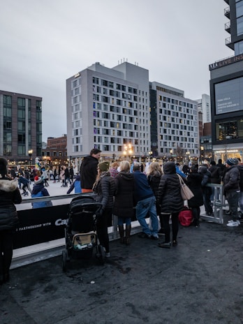 A bustling urban scene featuring an outdoor ice skating rink surrounded by modern buildings. People are ice skating and a group of onlookers are observing, some with children and strollers. The atmosphere suggests a winter setting with people dressed in warm clothing. The sky is overcast, adding to the chilly ambiance.