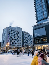People enjoy skating on an ice rink in an urban setting during the evening. Surrounding buildings feature modern architecture, and bright lights illuminate the area. Families and friends glide on the ice, some holding hands or assisting children.