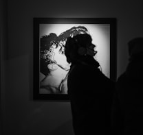 The image depicts a silhouette of a person wearing headphones, likely observing a framed artwork hanging on the wall. The artwork features an intricate black and white drawing of two people in an intimate pose, showing a close embrace or a kiss.