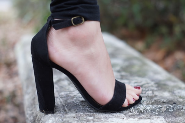 A close-up image of a person's foot wearing a black, high-heeled sandal with an ankle strap. The foot is resting on a textured stone surface, with a natural outdoor background that is blurred. The person has painted their toenails red.