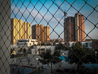 The image depicts tall apartment buildings in various shades of yellow and brown under a clear sky. A construction crane is visible near one of the taller buildings. A wire mesh fence is in the foreground, and below are residential houses and garden areas with visible trees and a swimming pool.