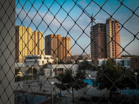 The image depicts tall apartment buildings in various shades of yellow and brown under a clear sky. A construction crane is visible near one of the taller buildings. A wire mesh fence is in the foreground, and below are residential houses and garden areas with visible trees and a swimming pool.