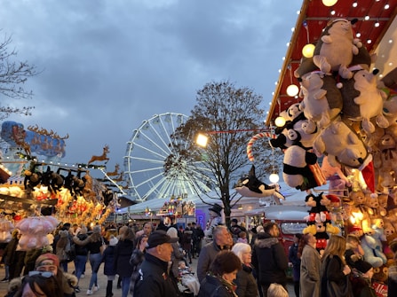 A lively fairground scene featuring a Ferris wheel in the background, numerous stalls selling various items, and many people walking around and enjoying the event. There are colorful stuffed toys hanging from a stall on the right side of the image and more toys and decorations on the left. The sky is overcast, suggesting it might be late afternoon or early evening.