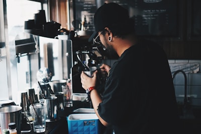 A barista is preparing a drink at an espresso machine in a coffee shop. The setting is dimly lit, with various coffee-making equipment visible, including glasses, a metal pitcher, and a blender. The barista is wearing a black cap and has a stylish earring.