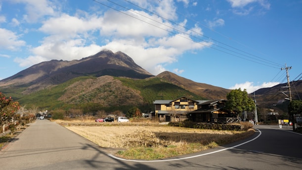 A rural landscape features a wide road curving around fields that are partially cultivated. In the background, a large mountain with a mix of green and brown vegetation dominates the scene. Several houses with traditional and modern architectural elements are situated alongside the fields. The sky is bright and partly cloudy, indicating fair weather.