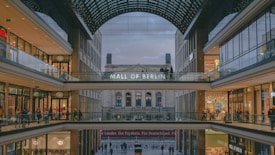 A large, modern shopping mall with multiple levels and a glass roof. The sign 'Mall of Berlin' is prominently displayed, visible to shoppers walking along the corridors. Numerous storefronts with visible brand signs and logos line the walkways. People are walking and shopping, creating a bustling atmosphere.