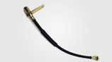 A flexible black rubber hose with metallic connectors at both ends, likely used for fluid or air transfer in mechanical systems. The connectors appear to be made of brass or a similar material and include a mounting bracket on one end.