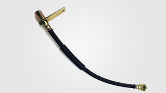 A flexible black rubber hose with metallic connectors at both ends, likely used for fluid or air transfer in mechanical systems. The connectors appear to be made of brass or a similar material and include a mounting bracket on one end.