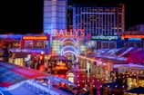 A brightly lit urban scene featuring the entrance to Bally's Las Vegas, surrounded by various illuminated shop signs including Giordano's, Wahlburgers, and Redneck Riviera. The street is bustling with people and rich with neon lights and colorful signage, creating a lively nighttime atmosphere.