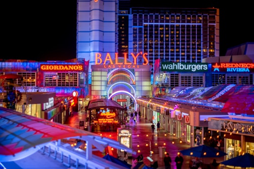 A brightly lit urban scene featuring the entrance to Bally's Las Vegas, surrounded by various illuminated shop signs including Giordano's, Wahlburgers, and Redneck Riviera. The street is bustling with people and rich with neon lights and colorful signage, creating a lively nighttime atmosphere.
