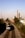 A freight train travels along a double railway track, heading towards an urban area. Tall buildings, including a prominent smokestack with the word 'AWW', dominate the skyline in the background. The scene is set under a clear blue sky with a mix of urban and natural landscape.
