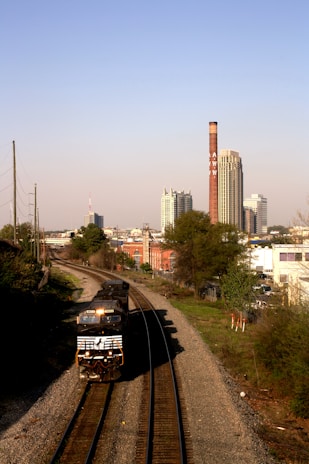 A freight train travels along a double railway track, heading towards an urban area. Tall buildings, including a prominent smokestack with the word 'AWW', dominate the skyline in the background. The scene is set under a clear blue sky with a mix of urban and natural landscape.