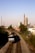 A freight train travels along a double railway track, heading towards an urban area. Tall buildings, including a prominent smokestack with the word 'AWW', dominate the skyline in the background. The scene is set under a clear blue sky with a mix of urban and natural landscape.