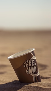 A disposable coffee cup is lying on its side on a beach with sand. The cup features text designs and branding, and the background shows a clear sky with a horizon in the distance.