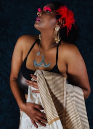 A person adorned with elaborate accessories, including prominent red feathers in their hair and bold red eyelashes, gazes upward. They wear a black top with a distinctive metallic necklace and large dangling earrings, while holding a patterned shawl draped elegantly over one shoulder. The background is a dark hue, contrasting with the vibrant elements.