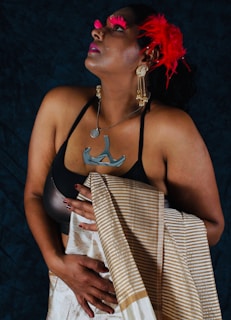 A person adorned with elaborate accessories, including prominent red feathers in their hair and bold red eyelashes, gazes upward. They wear a black top with a distinctive metallic necklace and large dangling earrings, while holding a patterned shawl draped elegantly over one shoulder. The background is a dark hue, contrasting with the vibrant elements.