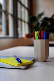 A neatly arranged desk with a yellow notepad and a blue pen resting on top. Nearby, a cup holds various colored markers. In the background, a window allows soft, natural light to fill the room, partially illuminating a plant.
