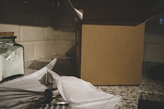 A kitchen counter scene with a partially opened cardboard box on the right, a glass jar filled with flour on the left, and a whisk partially wrapped in white paper. The countertop appears to be made of speckled granite, and white tile backsplashes are visible.