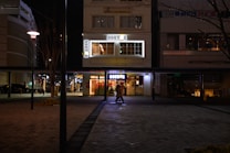 A nighttime street scene featuring a brightly illuminated caf&eacute; with the signage 'DONUT'. Two people are walking in front of the caf&eacute;, and the street is lined with modern buildings and a few trees without leaves. The area is lit by street lamps, creating a moody ambiance.