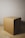 A plain cardboard box is placed on a wooden surface. The box has a simple, unadorned design with tape on its top sides. The lighting casts a shadow to the right, highlighting the texture and color of the cardboard.