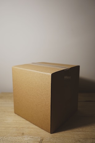 A plain cardboard box is placed on a wooden surface. The box has a simple, unadorned design with tape on its top sides. The lighting casts a shadow to the right, highlighting the texture and color of the cardboard.