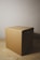 A plain cardboard box is placed on a wooden surface. The box has a simple, unadorned design with tape on its top sides. The lighting casts a shadow to the right, highlighting the texture and color of the cardboard.