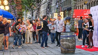 A bustling street scene with a crowd of people engaging in various activities. There are festive lights hanging from the trees and several vendors with colorful umbrellas and illuminated signage. A large barrel is placed in the center, and a few people are posing in front of a promotional backdrop.