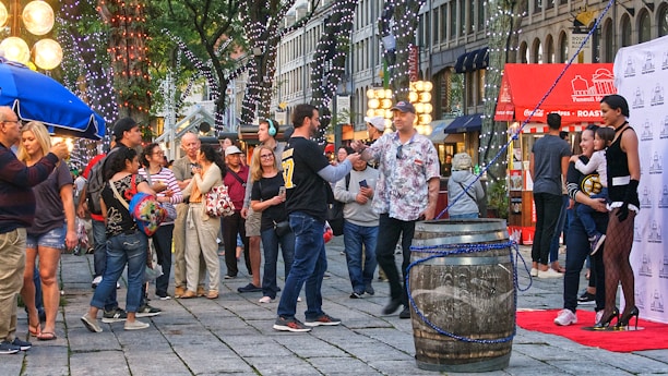 A bustling street scene with a crowd of people engaging in various activities. There are festive lights hanging from the trees and several vendors with colorful umbrellas and illuminated signage. A large barrel is placed in the center, and a few people are posing in front of a promotional backdrop.