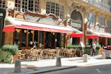 A charming street scene featuring a Parisian bistro with an ornate exterior and red awnings. Wooden chairs and tables are set outside for dining, and a person is seen organizing the setup. Another couple is seen standing nearby, adding to the casual and bustling atmosphere. The facade includes elegant architectural details and large windows.