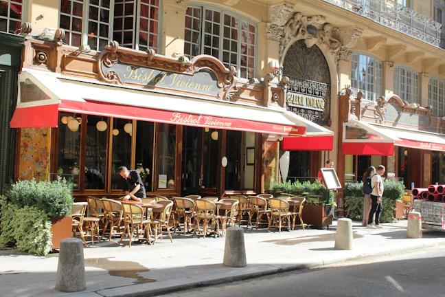 A charming street scene featuring a Parisian bistro with an ornate exterior and red awnings. Wooden chairs and tables are set outside for dining, and a person is seen organizing the setup. Another couple is seen standing nearby, adding to the casual and bustling atmosphere. The facade includes elegant architectural details and large windows.