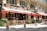A charming street scene featuring a Parisian bistro with an ornate exterior and red awnings. Wooden chairs and tables are set outside for dining, and a person is seen organizing the setup. Another couple is seen standing nearby, adding to the casual and bustling atmosphere. The facade includes elegant architectural details and large windows.