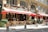 A charming street scene featuring a Parisian bistro with an ornate exterior and red awnings. Wooden chairs and tables are set outside for dining, and a person is seen organizing the setup. Another couple is seen standing nearby, adding to the casual and bustling atmosphere. The facade includes elegant architectural details and large windows.