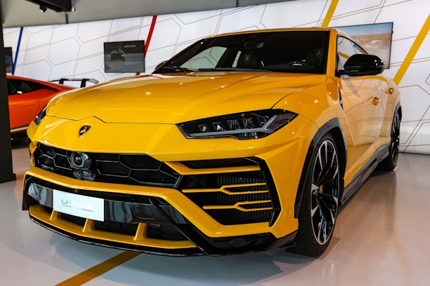 A sleek, yellow luxury SUV with an aggressive front grille and stylish headlights is displayed in a showroom. The car has a high-gloss finish and black trim accents. The background includes geometric patterns and another vehicle in orange.